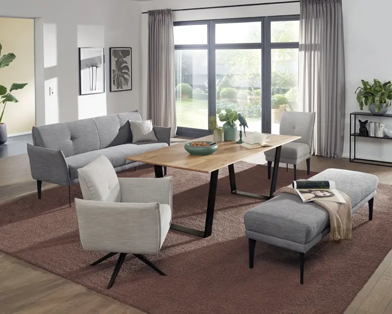 Wohnzimmer mit Sofa, zwei Sesseln, Hocker und Holztisch vor großer Fensterfront
